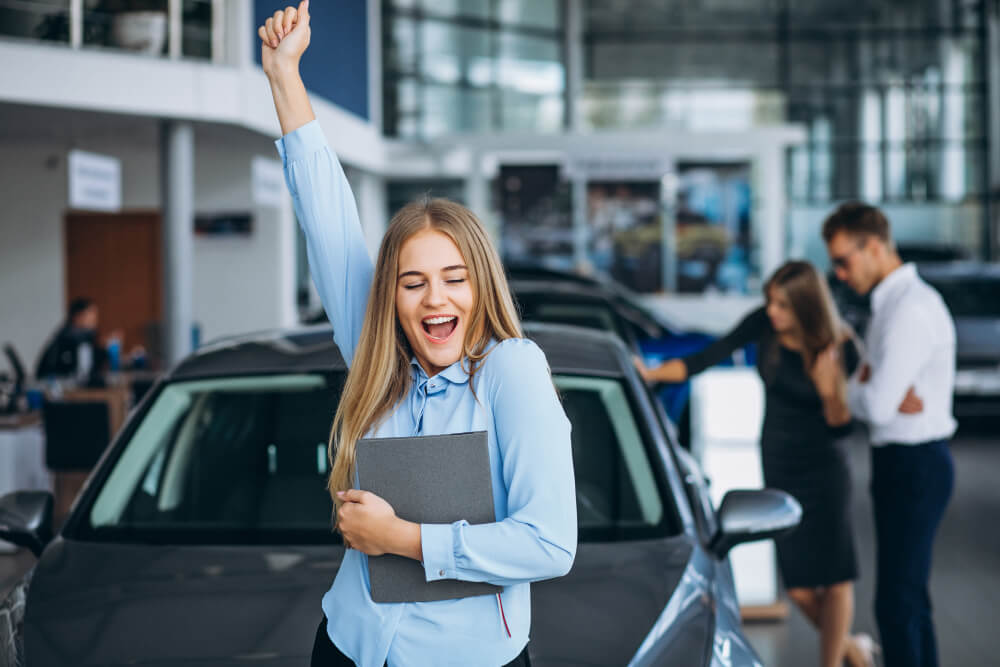 mulher vendedora de carros comemorando o sucesso nas vendas, com casal de clientes ao fundo ao lado do carro comprado