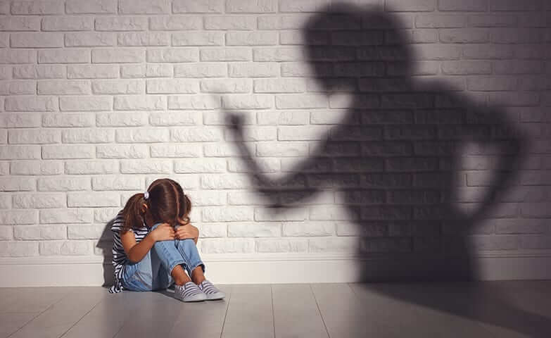 Criança acuada sentada no chão com a cabeça entre os joelhos. Abraçando os joelhos. Em frente à uma parede de tijolos brancos. Sombra de uma mulher na parede apontando o dedo para a menina.